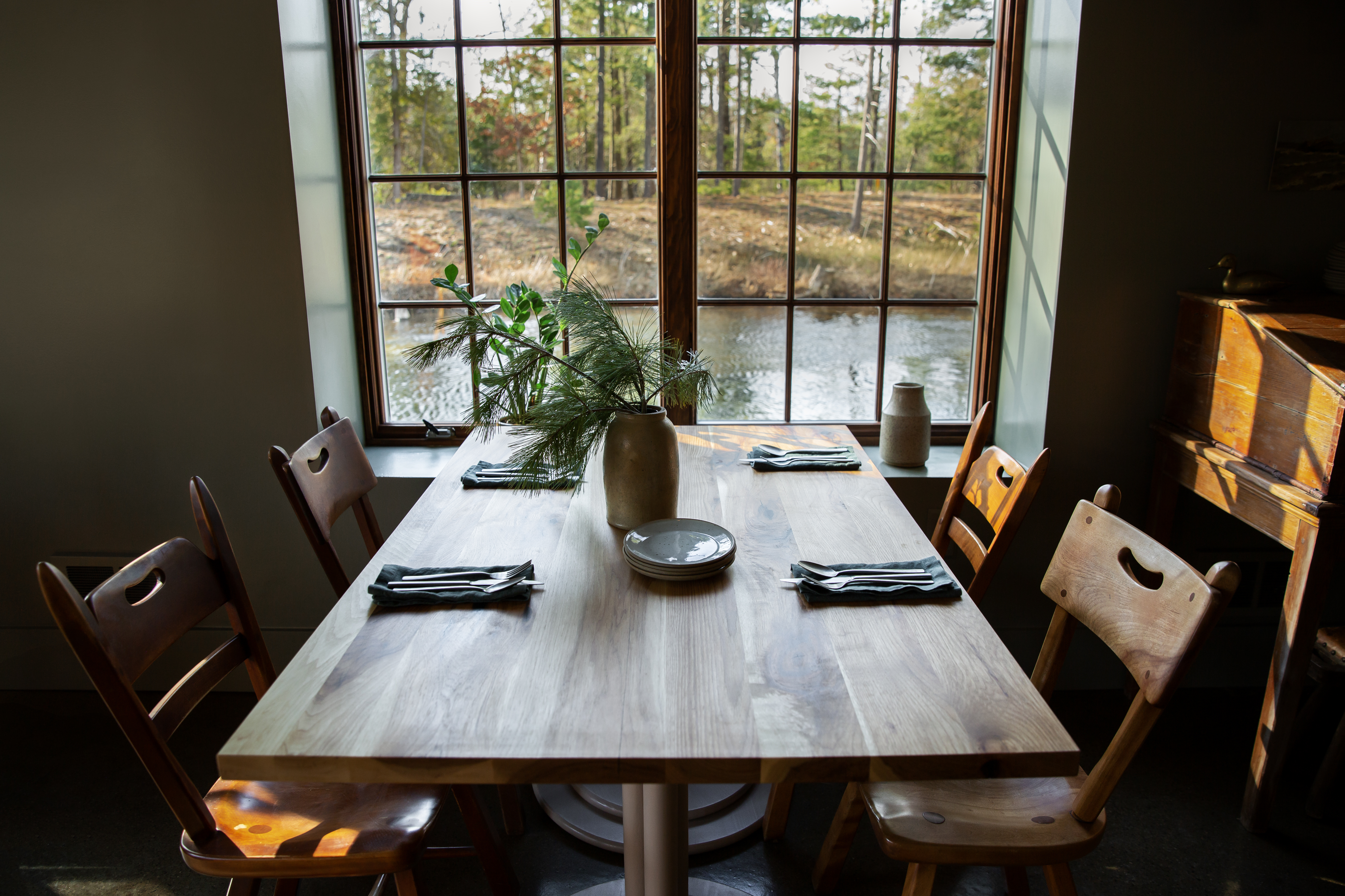 A wooden table surrounded by chairs with table settings on top, a vase, sat next to a window facing water.