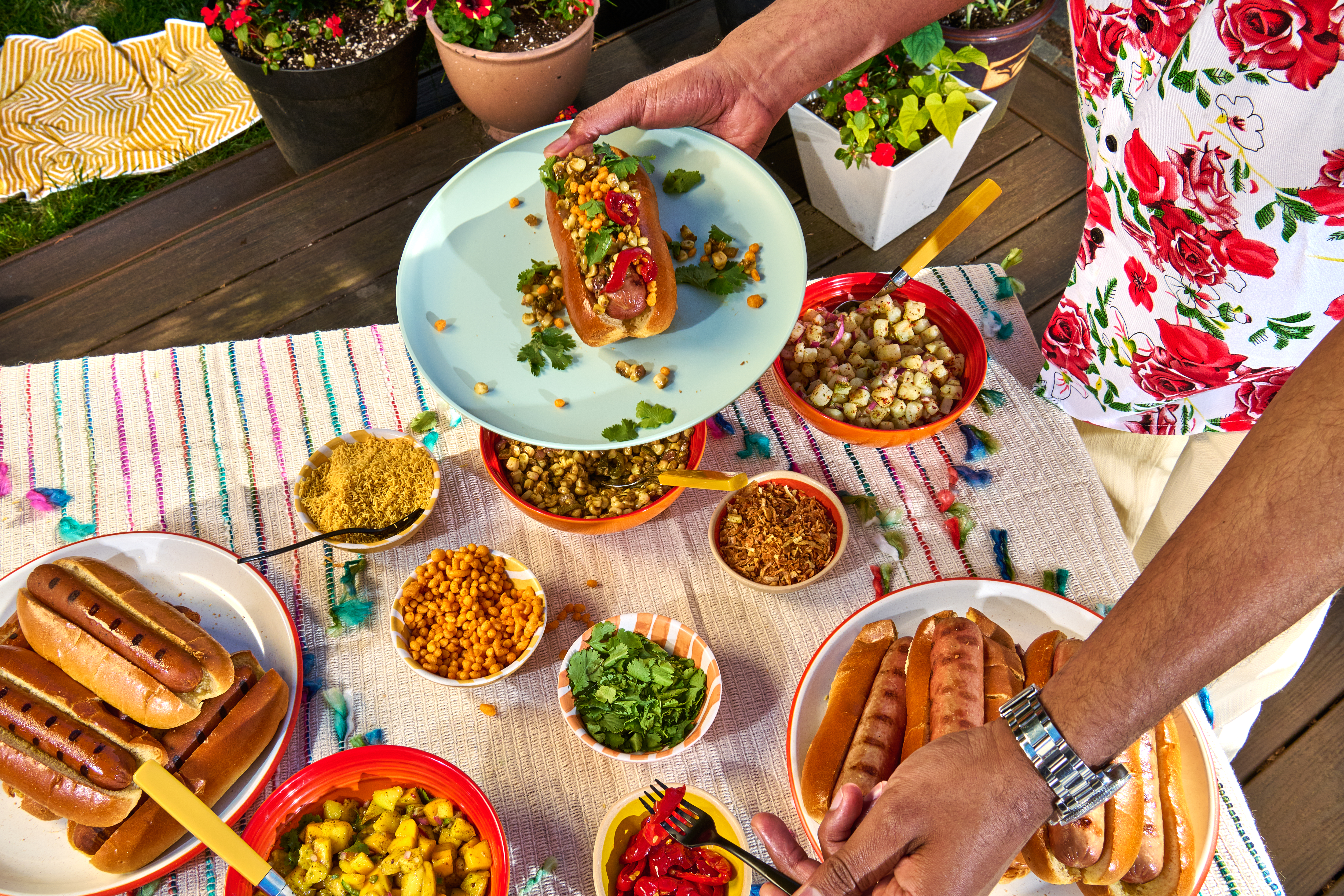 A person holds a plate with a chaat dog over a table set with platters of hot dogs in buns and bowls of chaat.