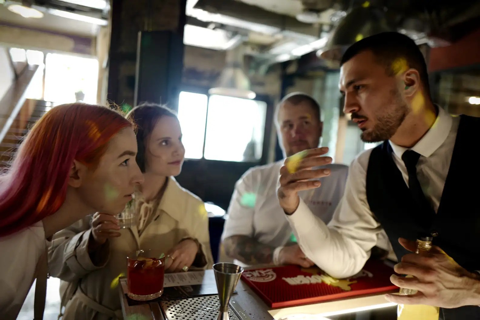 A group of four people — two women and two men, including one wearing a vest and tie — lean forward over a bar in discussion. 