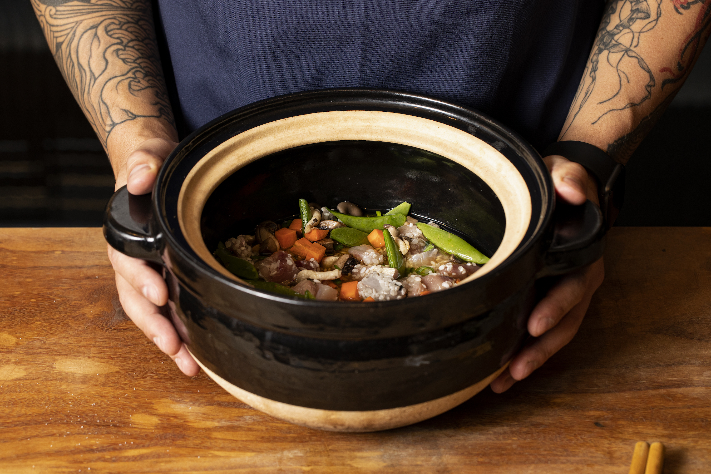 A chef holds a donabe full of kamamaeshi on a wooden countertop. 