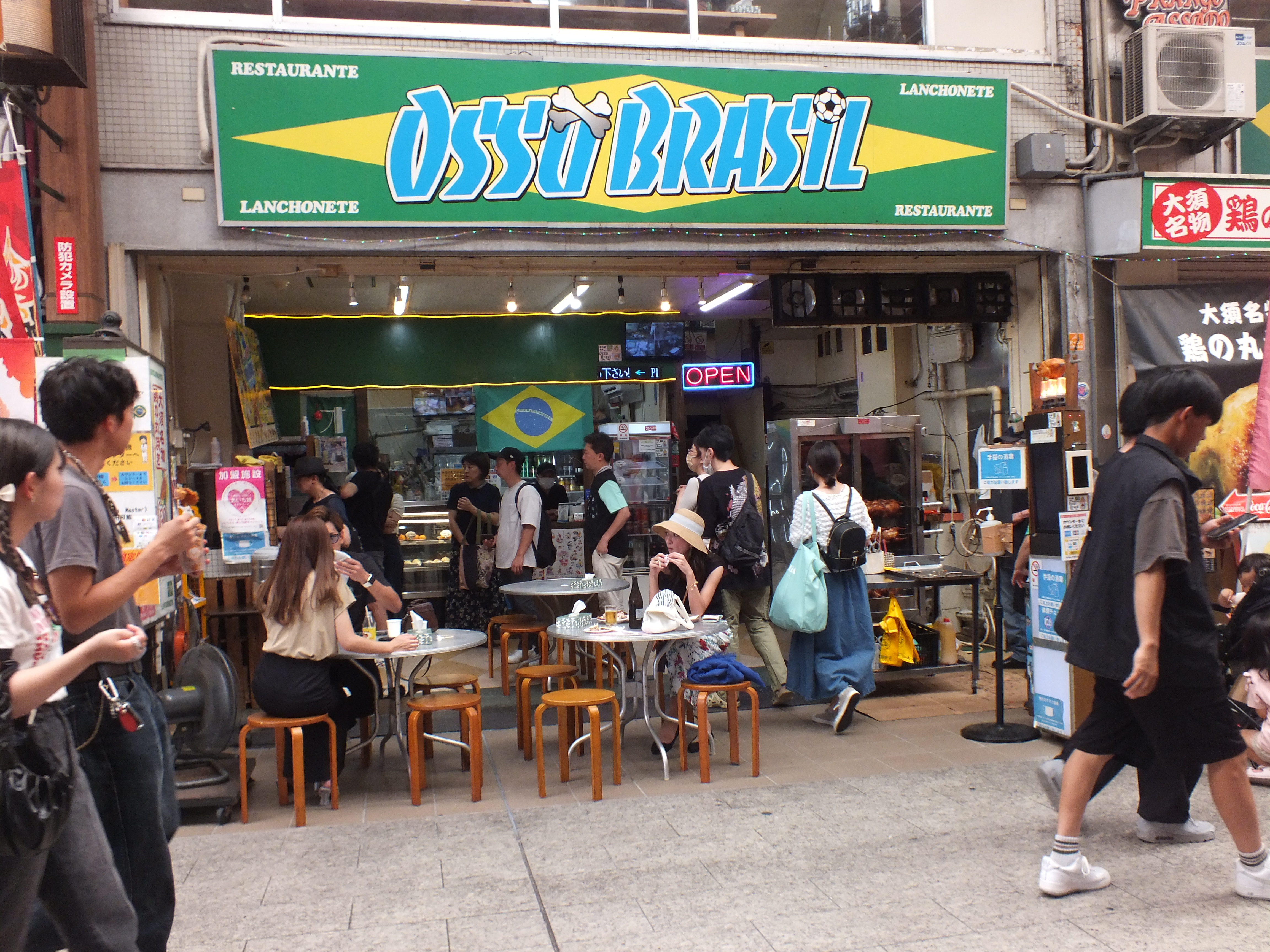 Customers dine at and walk by a restaurant open to the sidewalk, with a sign reading Osso Brasil over a Brazilian flag.