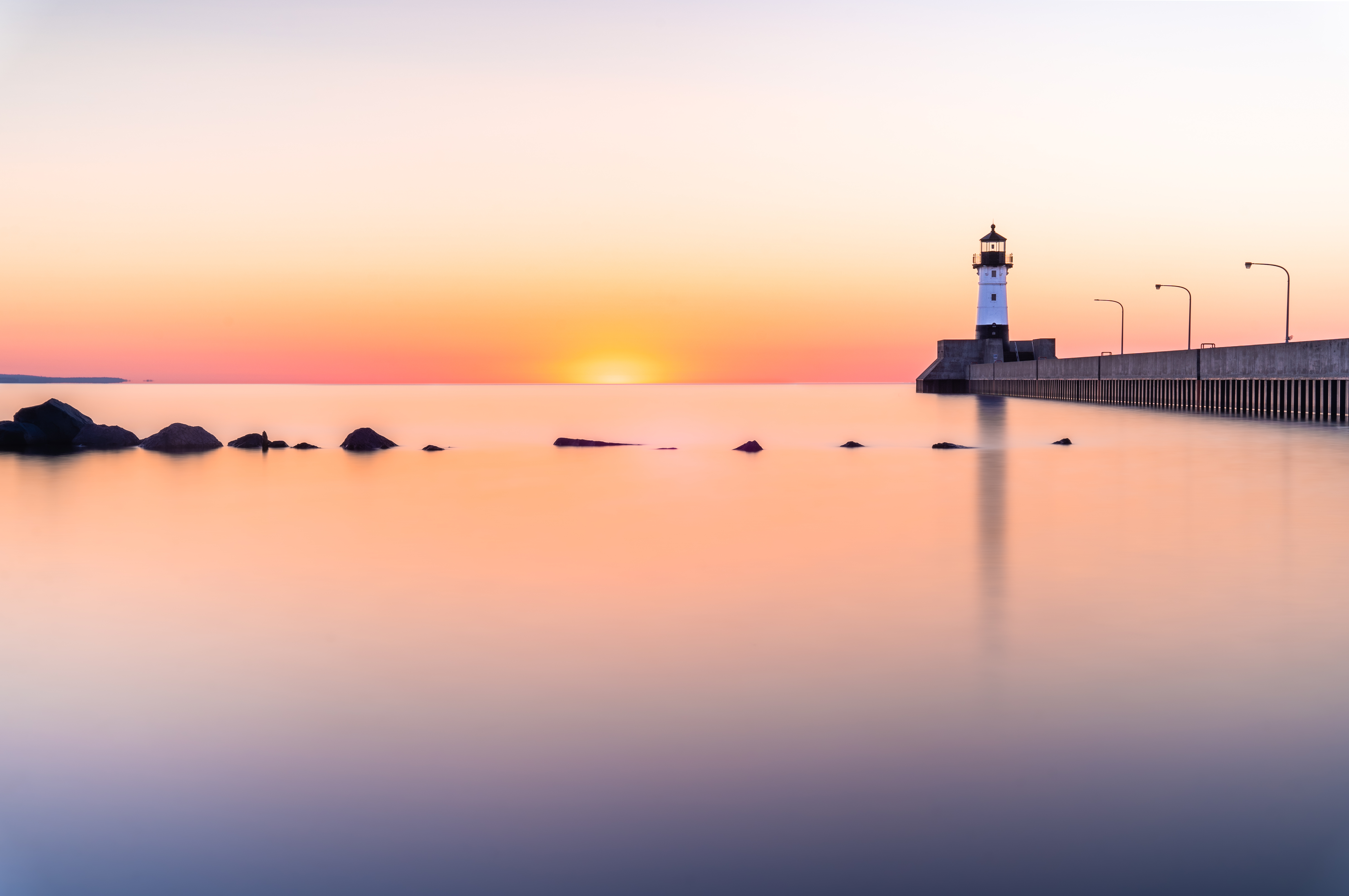 A sunset above a placed lake with a row of rocks in the middleground and a pier and lighthouse in the background. 