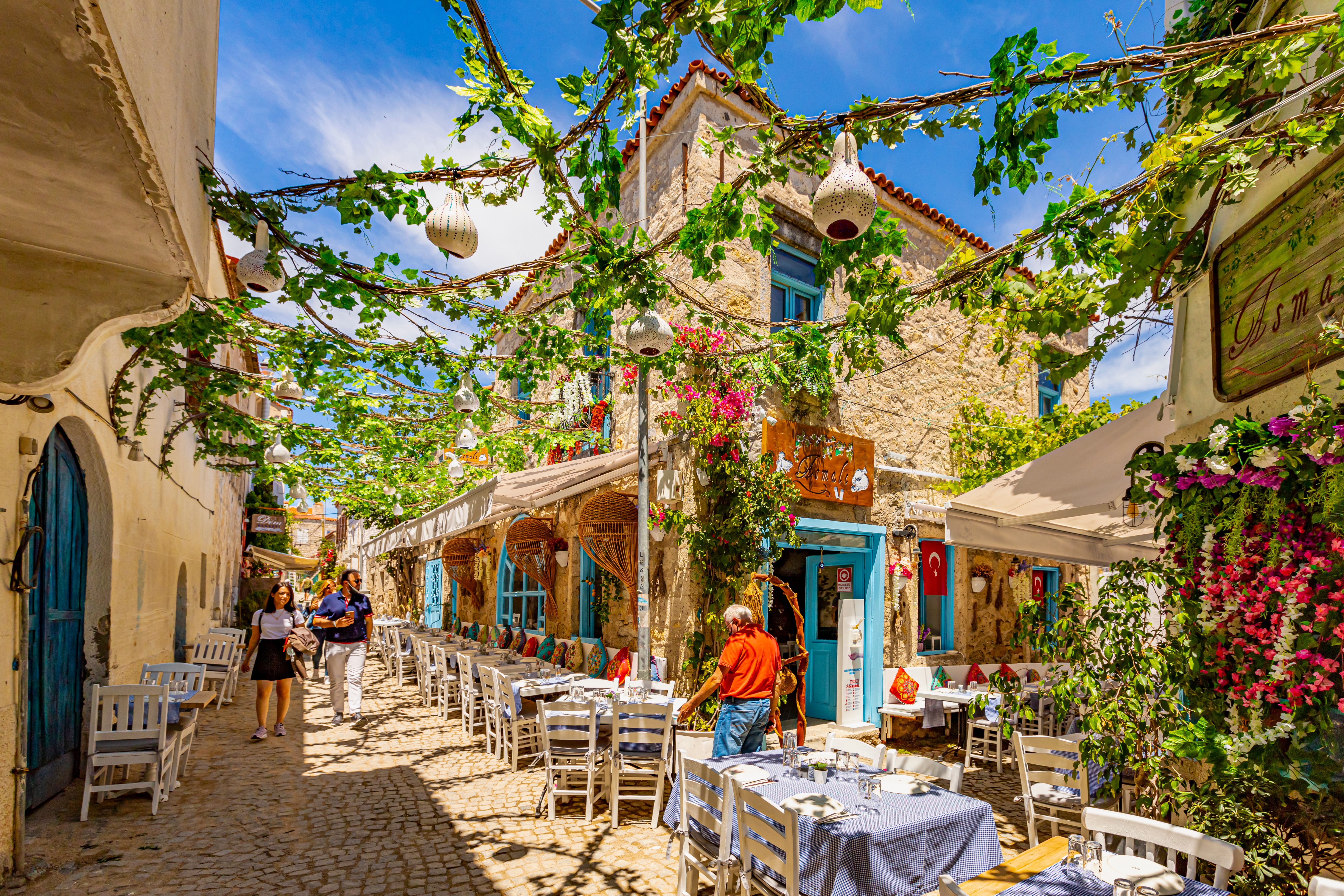 Pedetrians wander stone streets beneath greenery and brightly painted shutters, while a proprietor sets up outdoor seating at a restaurant. 