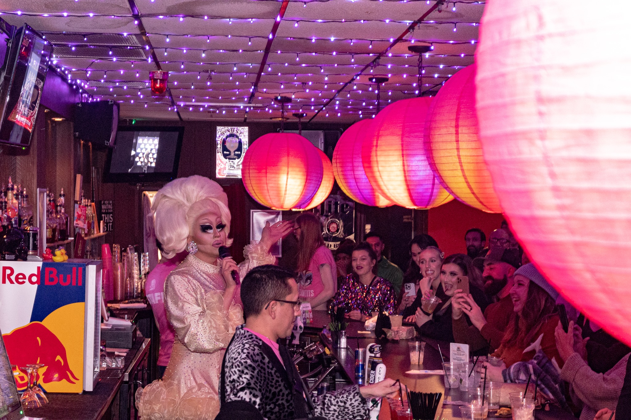 Trixie Mattel, a drag performer with giant blonde wig, holds the microphone among a seated crowd in a bar with pink and red lanterns.
