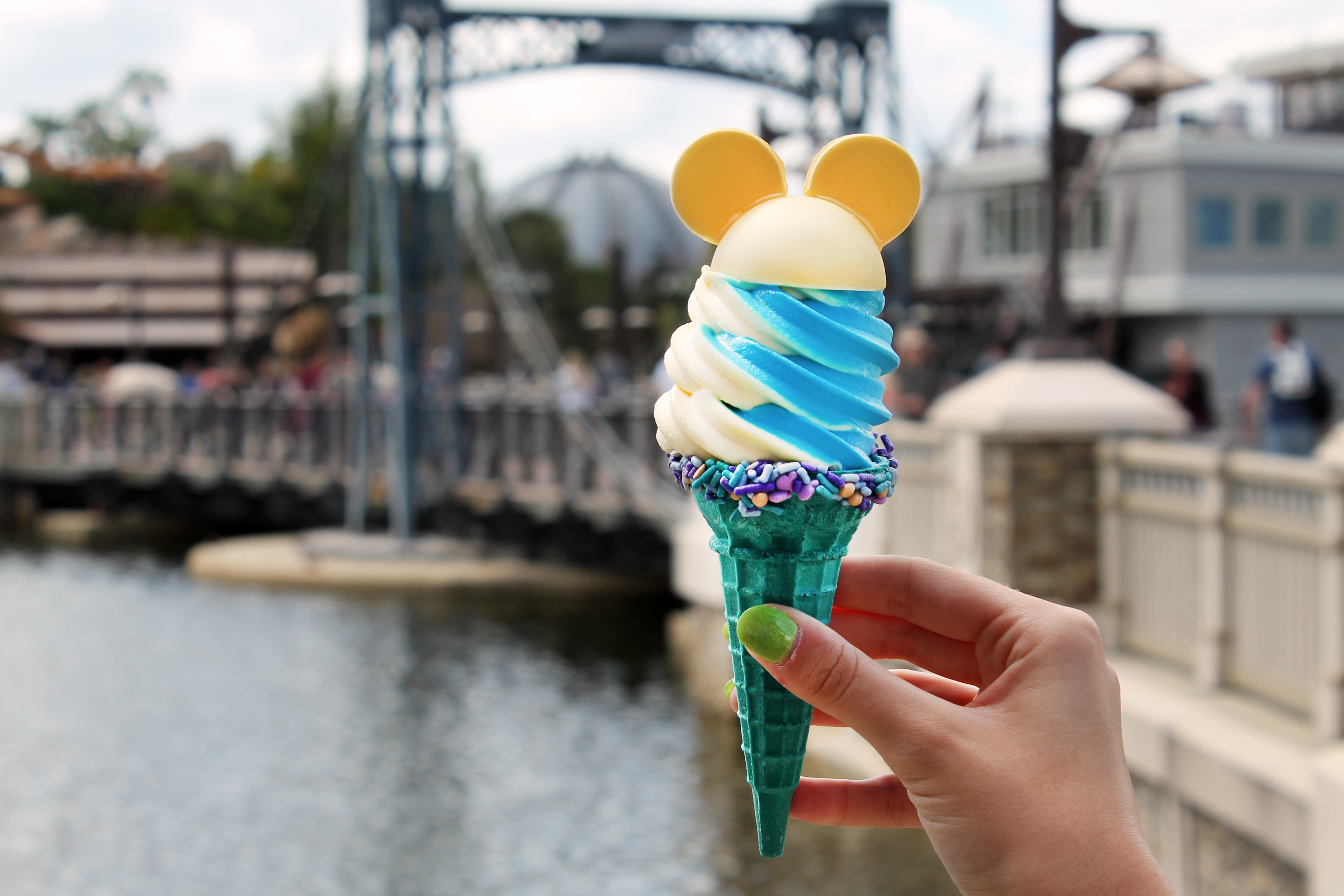 A teal-colored ice cream cone of white and blue soft serve swirled with a rim of sprinkles around the cone and cookies in the shape of Mickey Mouse ears on top