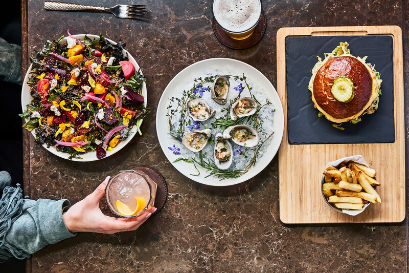 Bird’s-eye-view of an oyster, burger, and cocktail spread sitting atop a marble counter.